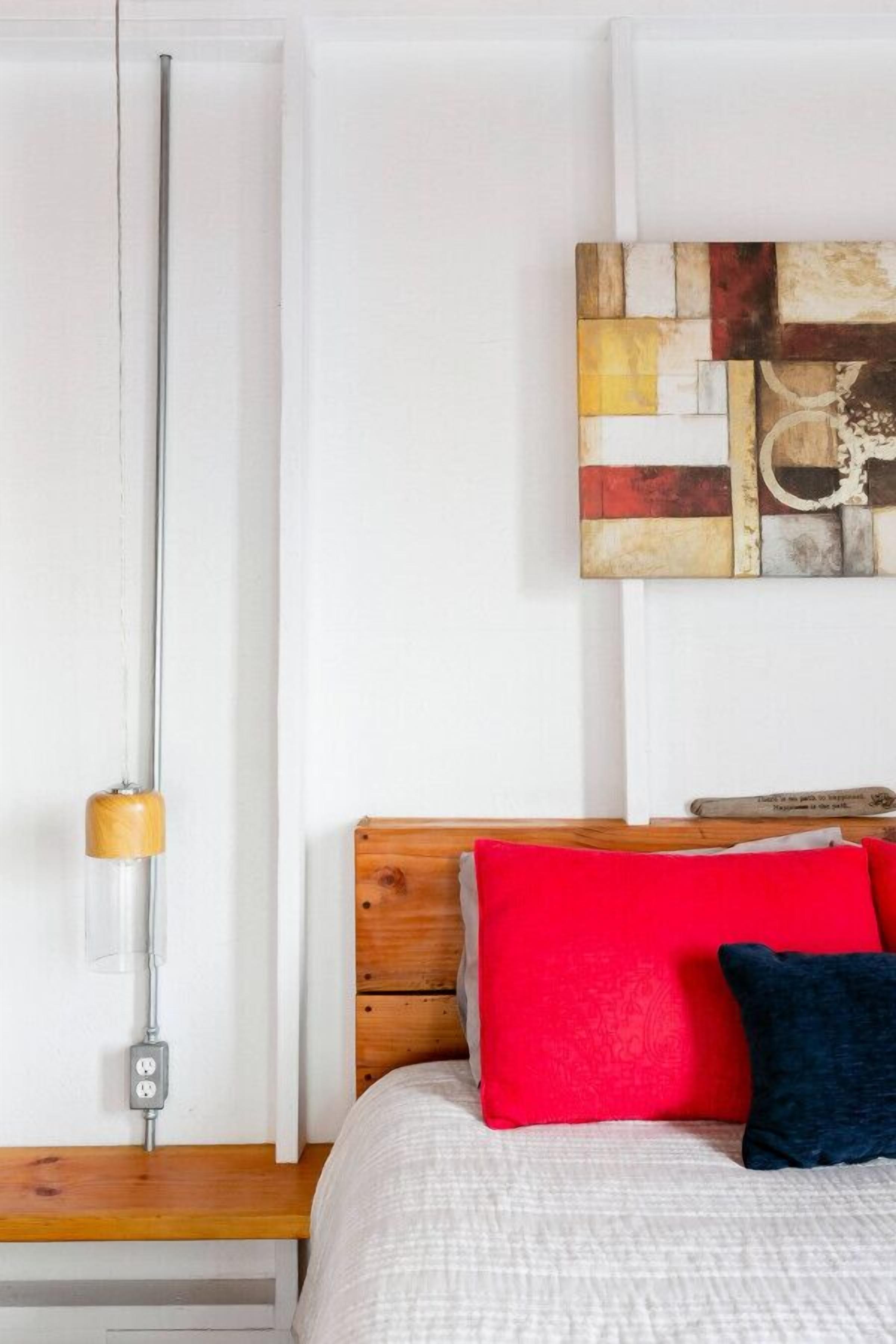 Une chambre à coucher avec une tête de lit en bois, un lit recouvert d’une literie blanche et rouge, une œuvre d’art abstraite accrochée au mur et une table de chevet avec une lampe.