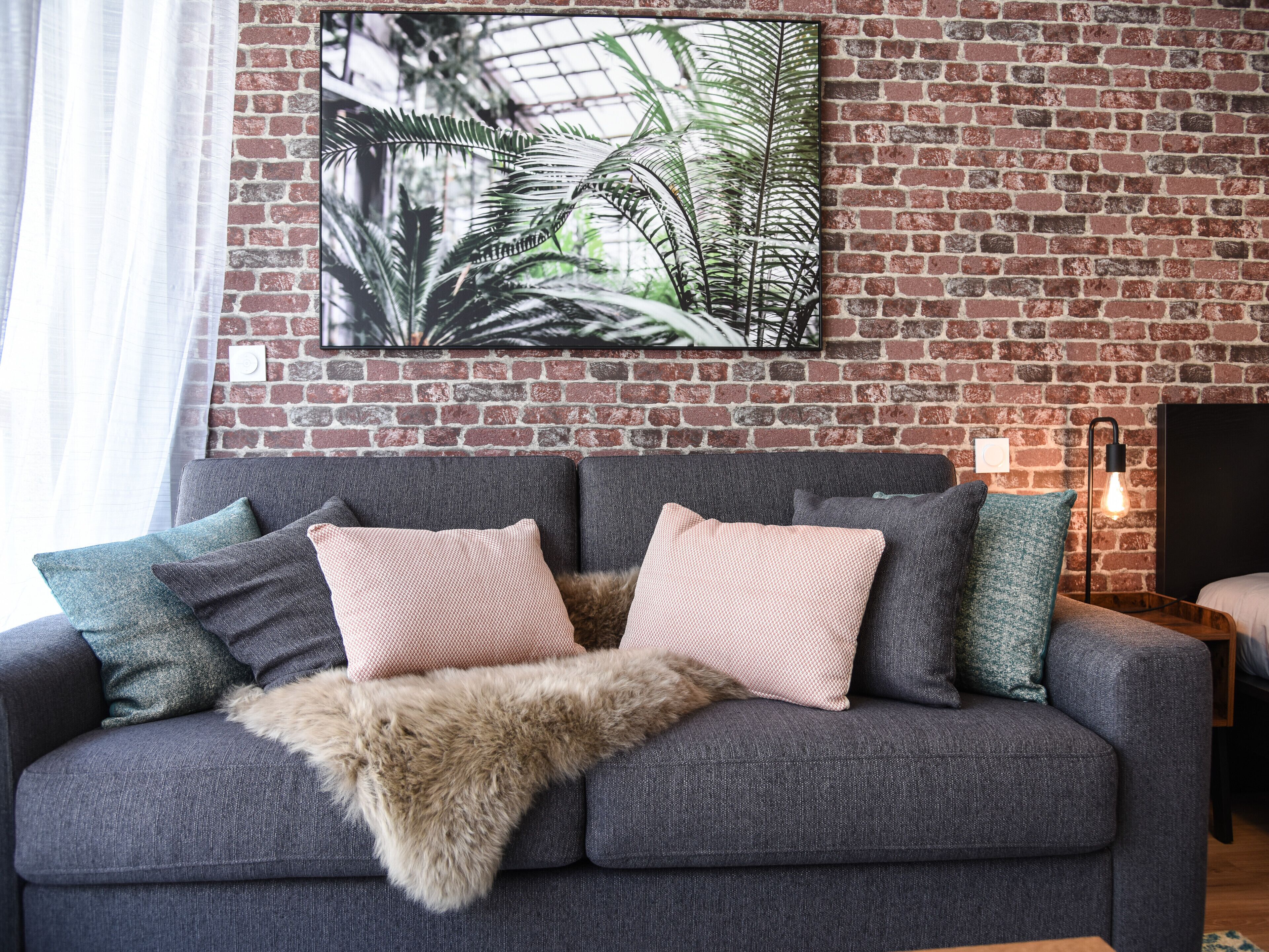 A grey sofa with two pink pillows and a fur throw, against a brick wall with a framed plant artwork.