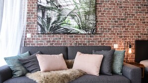 Ein graues Sofa mit zwei rosafarbenen Kissen und einer Felldecke vor einer Ziegelwand, an der ein Bild mit einer Pflanze hängt.