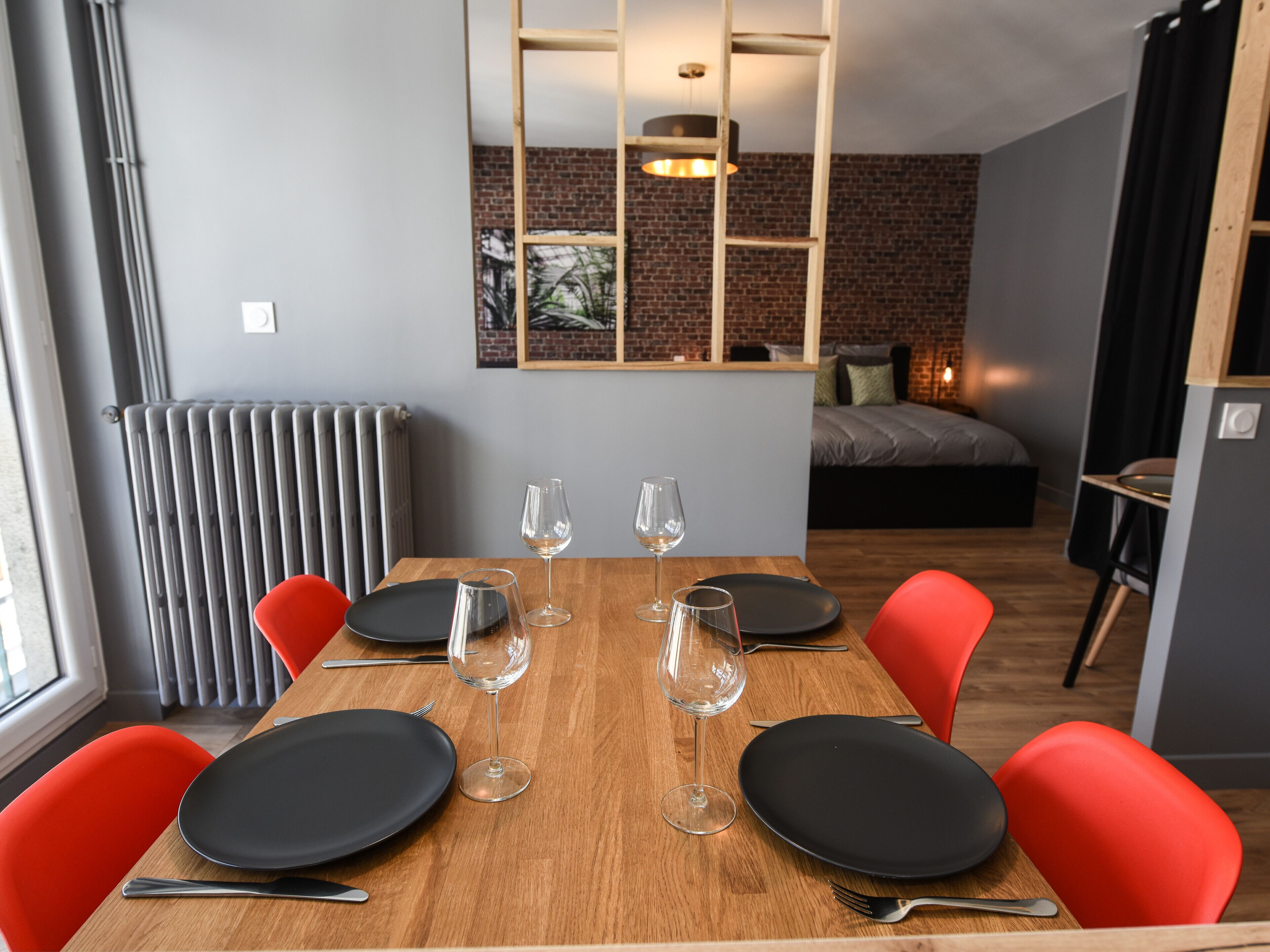 A dining area with a wooden table set for four, surrounded by red chairs. There are four wine glasses and plates on the table. A brick wall and a bed with a gray mattress and pillows are visible in the background.