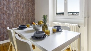 A dining table with a white tablecloth, surrounded by white chairs. There is a fruit bowl with a pineapple and bananas, a glass of orange juice, and a bowl of cereal. - Appartement Bourbon - Welkeys (Bayonne)