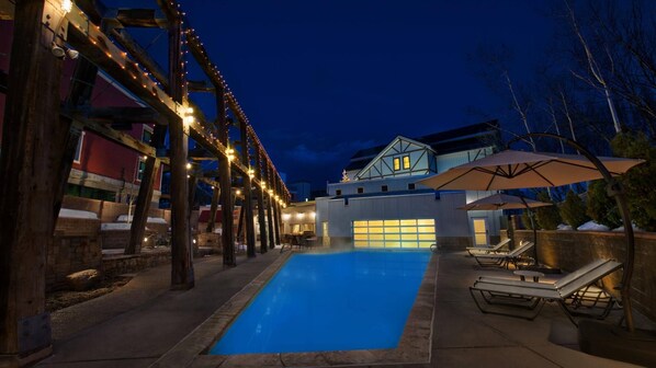 A swimming pool with lounge chairs and umbrellas, surrounded by a wooden deck and string lights.