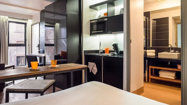 A modern hotel room with a wooden desk, a bed, a kitchenette, and a bathroom with a large mirror. - Vieille Bourse - Studio close to stations and old city - Welkeys (Lille)