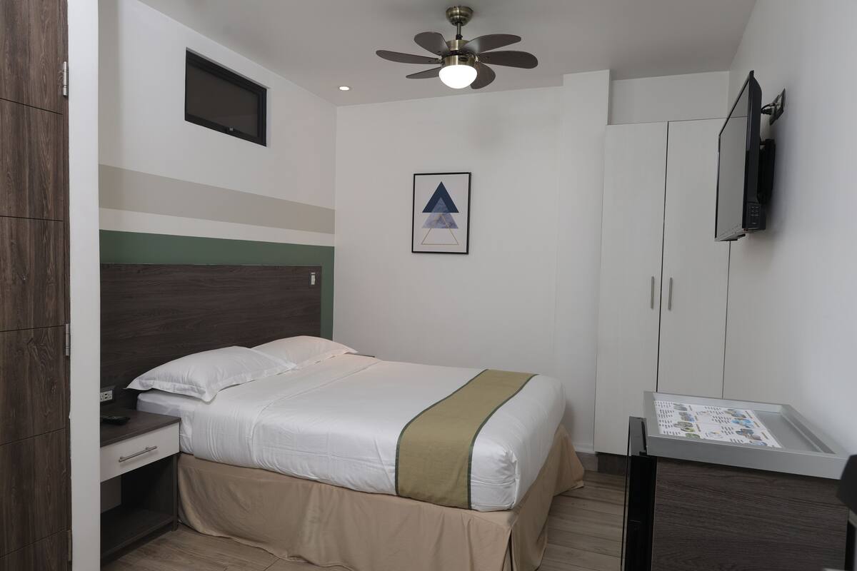 A hotel room with a bed, a desk, a chair, a TV, and a ceiling fan.