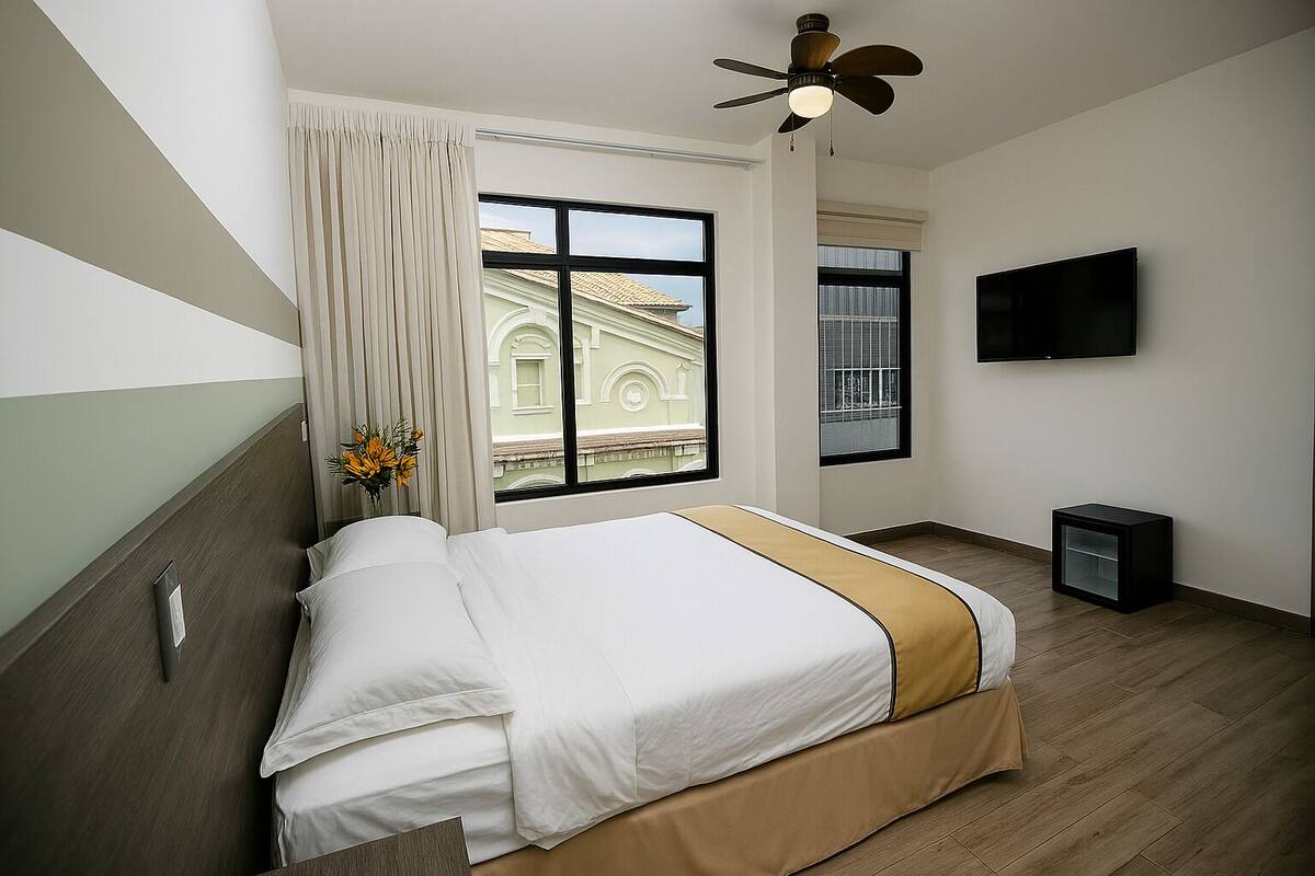 A hotel room with a bed, a TV, a ceiling fan, and a window with a view of a building.