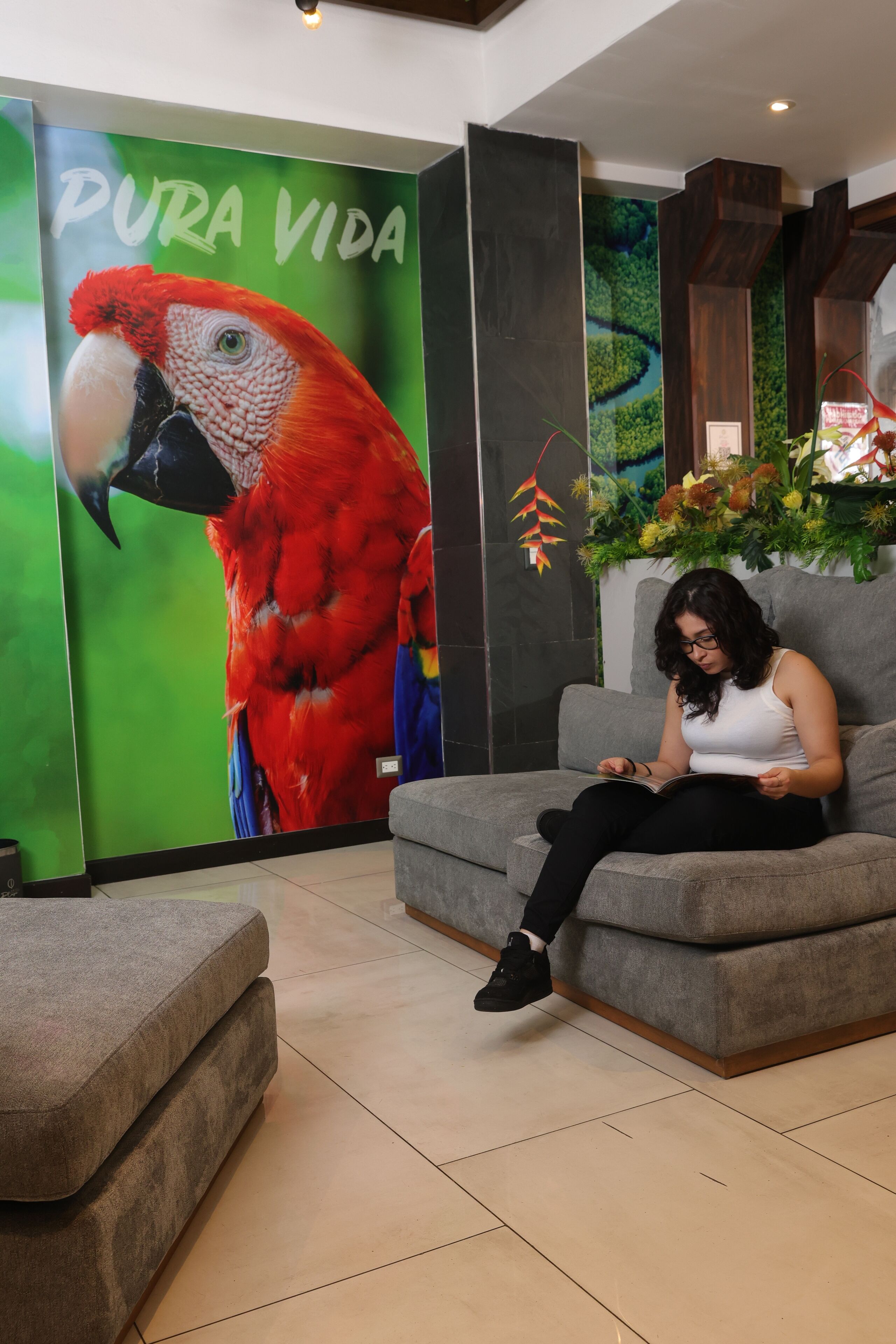 A woman sitting on a grey sofa in a lobby with a large parrot mural and a sign reading "PURA VIDA".
