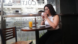 A person sitting at a table with a glass of orange juice and a white cup, in a cafe with a view of buildings through the window. - Hotel Cultura Plaza (San José)