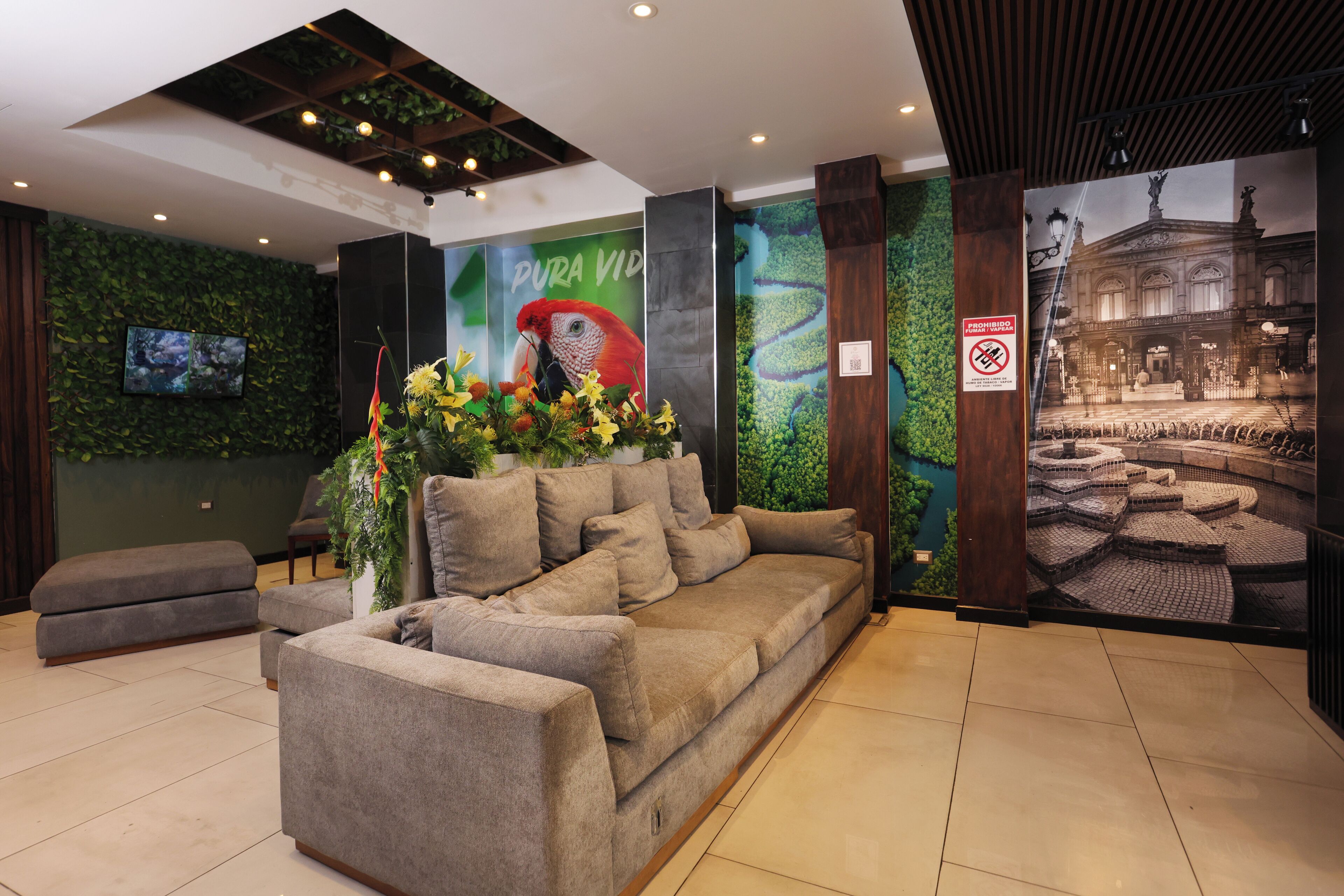 A lobby with a sofa, a television, a wall with greenery, and a poster of a building.