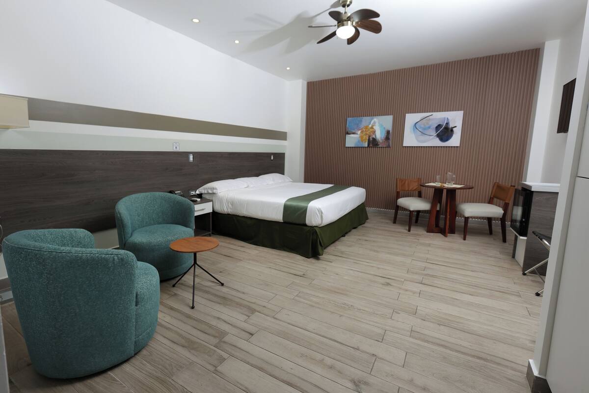 A hotel room with a bed, two teal armchairs, a small round table, a ceiling fan, and two paintings on the wall.