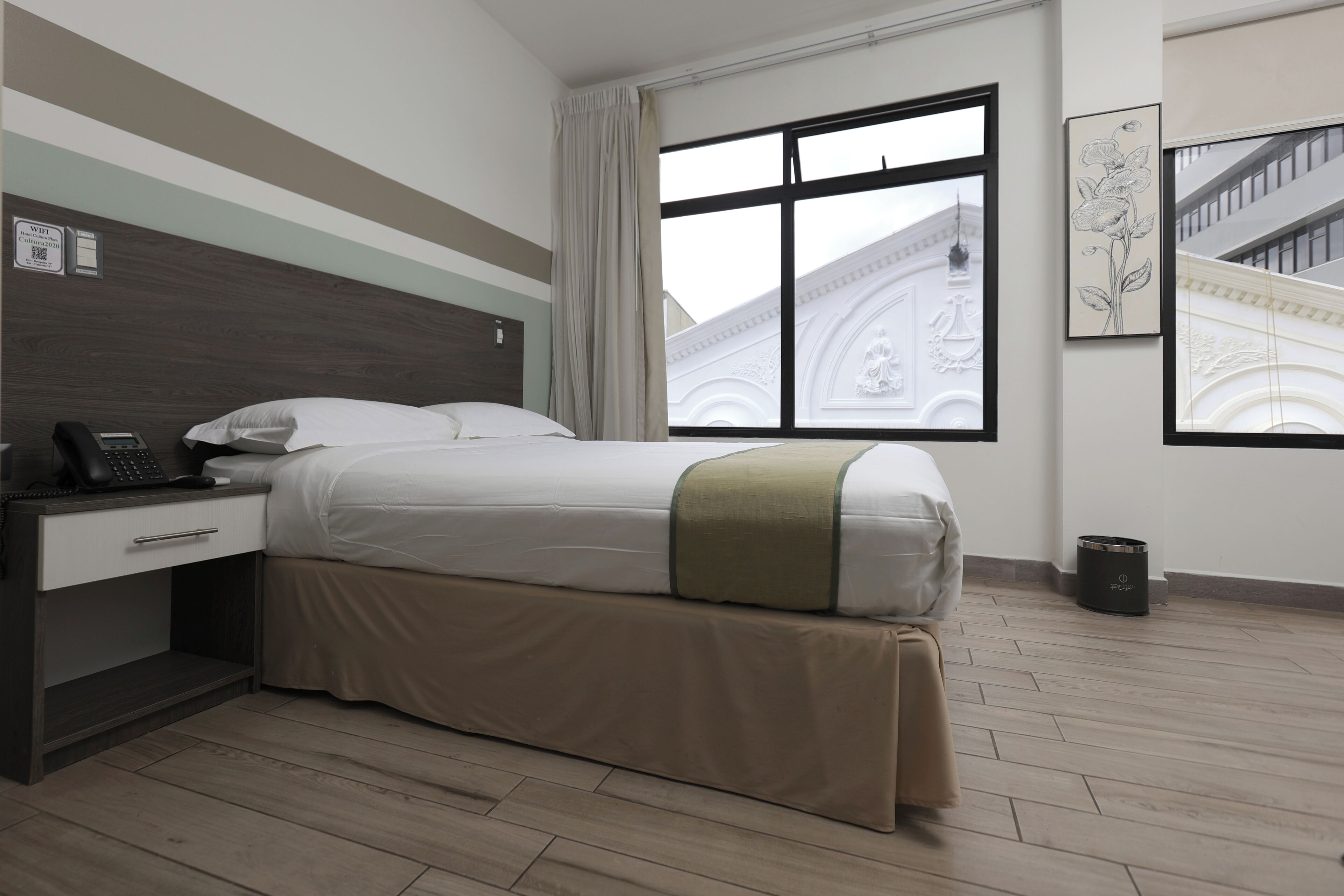 A hotel room with a bed, a desk with a phone, a trash bin, and a window with a view of a building.