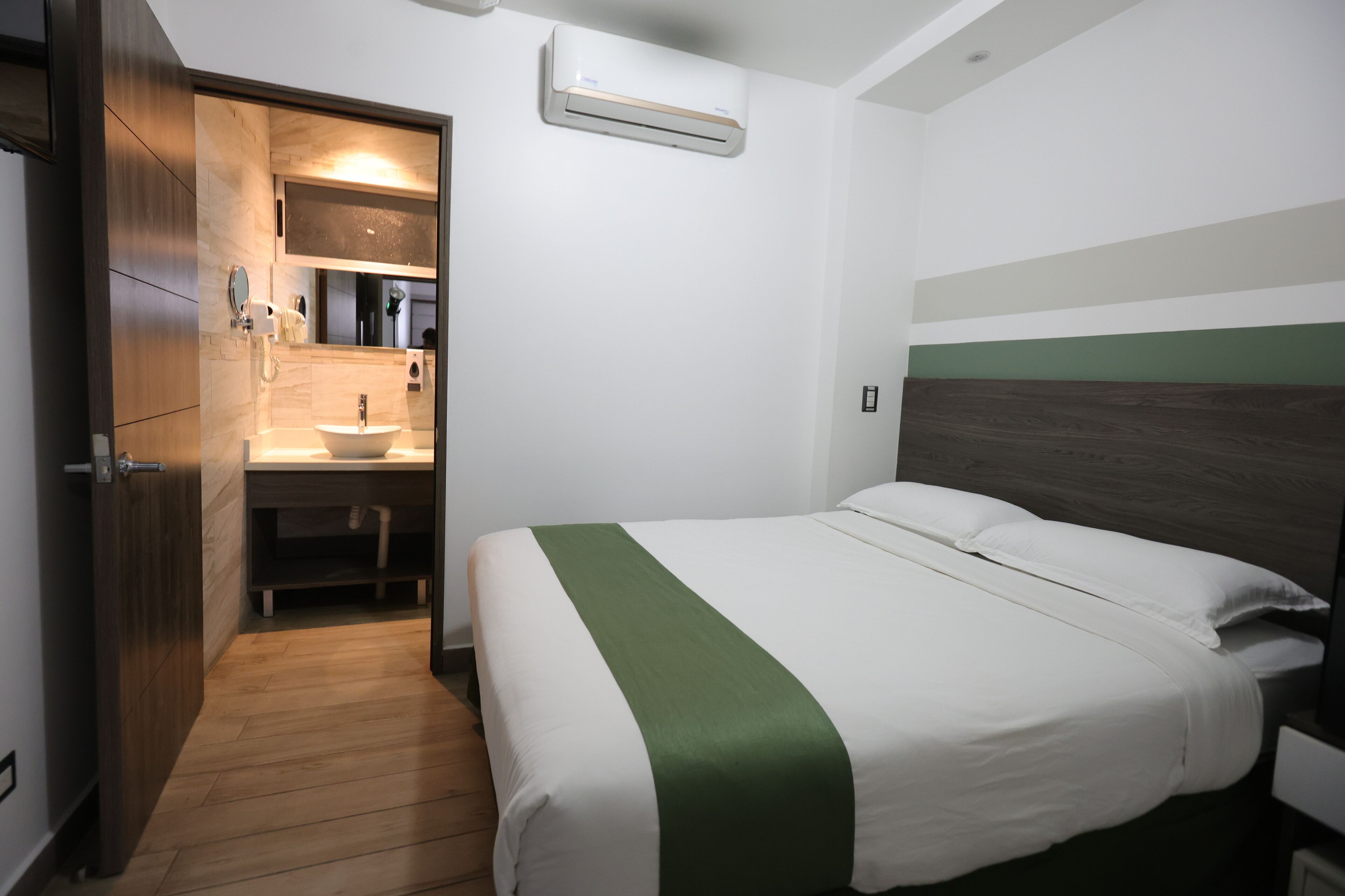 Quarto de hotel com piso de madeira, cama com roupa de cama branca e verde, mesa de cabeceira, espelho e banheiro com pia e espelho.