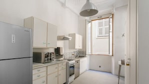 A compact kitchen with white cabinetry, a stainless steel refrigerator, a built-in oven, and a window with blinds. - Appartement Verrière - Welkeys (Nice)