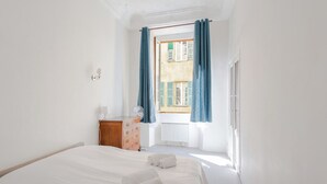 A bedroom with a large bed, a dresser, a radiator, and a window with blue curtains. - Appartement Verrière - Welkeys (Nice)