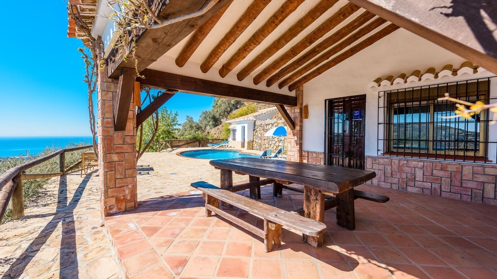 A covered patio with a wooden table and benches, a swimming pool, and a view of the sea.