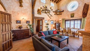Un vaste salon doté d’un grand canapé, un coin repas avec des meubles en bois et un intérieur rustique agrémenté de poutres apparentes et d’un lustre.