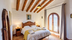 Une chambre avec un plafond en bois, une grande fenêtre avec des rideaux, un lit avec du linge de lit blanc et un tableau au mur.