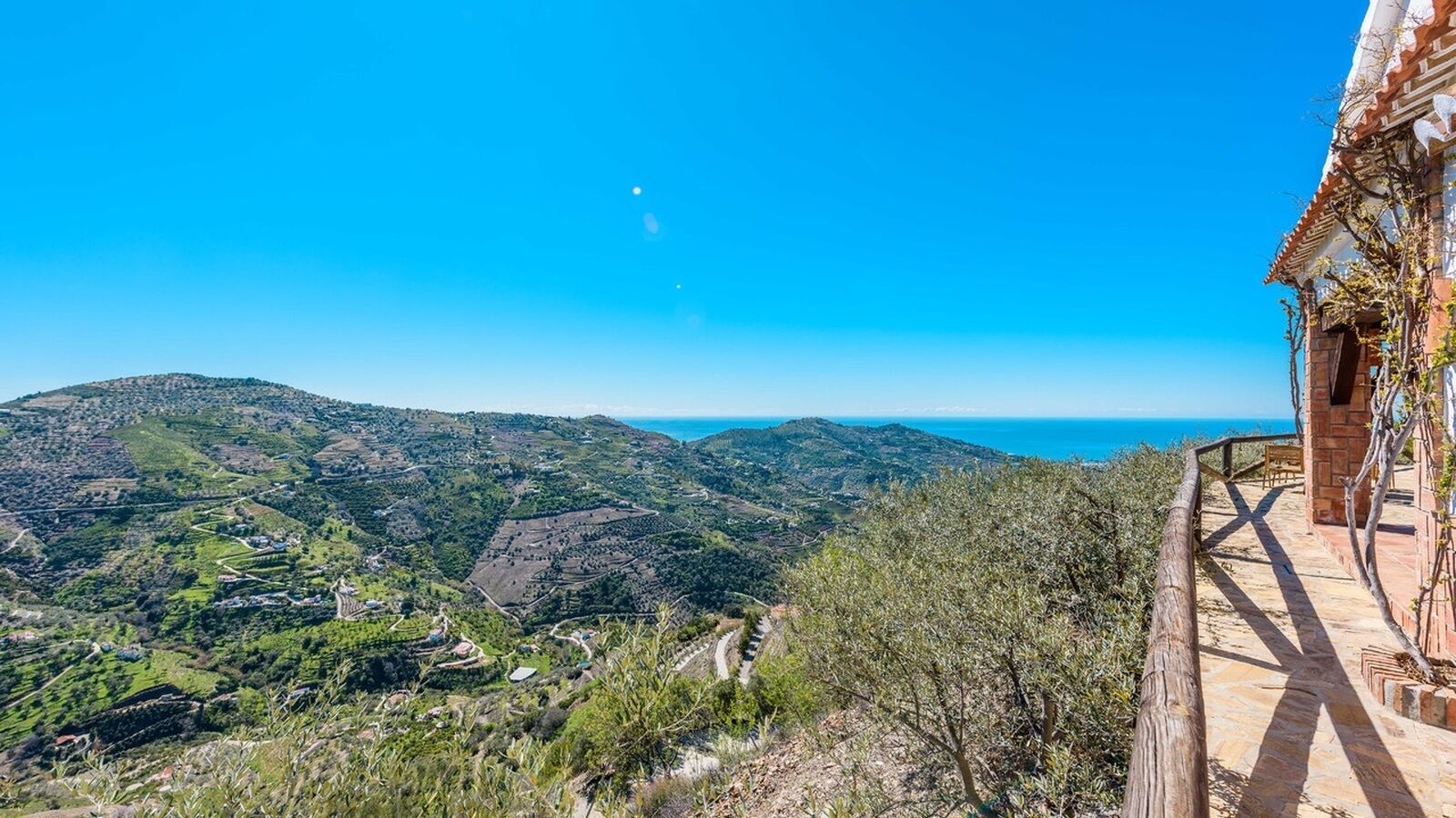 A hillside view with a clear sky, a distant body of water, and a building with a wooden balcony.