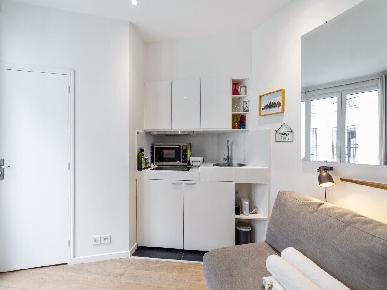 A compact kitchen with white cabinets, a microwave, and a sink. There's a grey sofa and a lamp in the living area.