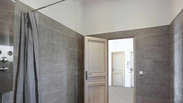 A modern bathroom with grey tiles, a shower curtain, and a wooden door leading to another room. - Appartement Carnot - Welkeys (Cannes)