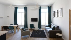 A modern living room with a dining area, a sofa, a TV mounted on the wall, and large windows with curtains. - Appartement Carnot - Welkeys (Cannes)
