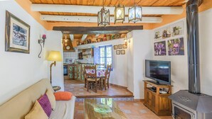 Un salón acogedor con sofá, televisión y chimenea. Hay una zona de comedor con sillas y mesa. La habitación tiene vigas de madera en el techo y suelo de baldosas.