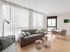 A modern living room with a sofa, a dining table with chairs, and a large window with sheer curtains.