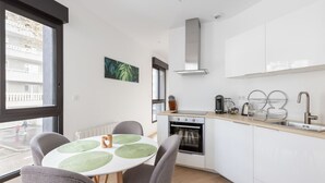 A modern kitchen with white cabinets, a stainless steel range hood, and a built-in oven. There is a dining table with four chairs and a window providing a view of the street. - Appartement Céladon - Welkeys (Lyon)