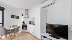 A modern kitchen with white cabinets, a dining area with a table and chairs, and a flat-screen TV on a white stand. - Appartement Céladon - Welkeys (Lyon)