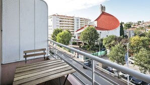 Ein Balkon mit einer Holzbank, einer Brüstung und Blick auf eine Straße mit Gebäuden und Bäumen.