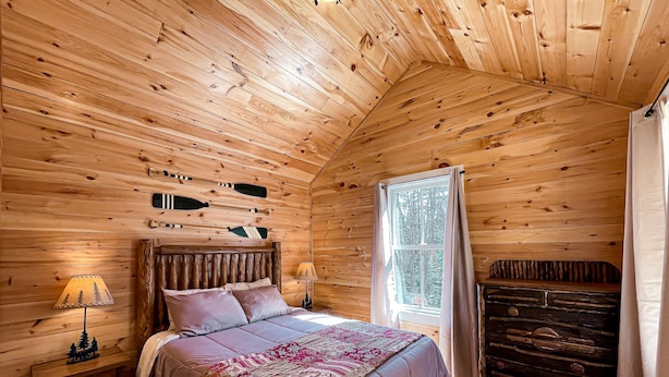 Un dormitorio acogedor tipo cabaña de madera, con cama, mesitas de noche, cómoda y ventana con cortinas.
