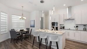 A modern kitchen with a central island, bar stools, a dining area with a table and chairs, and a large window with blinds. - Your Ultimate Luxury Getaway in the Providence Golf Community (Davenport)