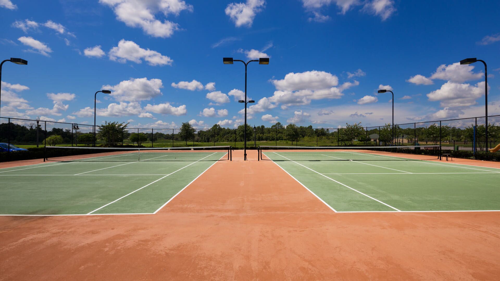 A tennis court with a clay surface, surrounded by a fence, and under clear skies.