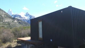 Une petite cabane aux couleurs sombres, dotée d’une terrasse en bois et d’une porte, se dressant sur un fond de paysage montagneux.