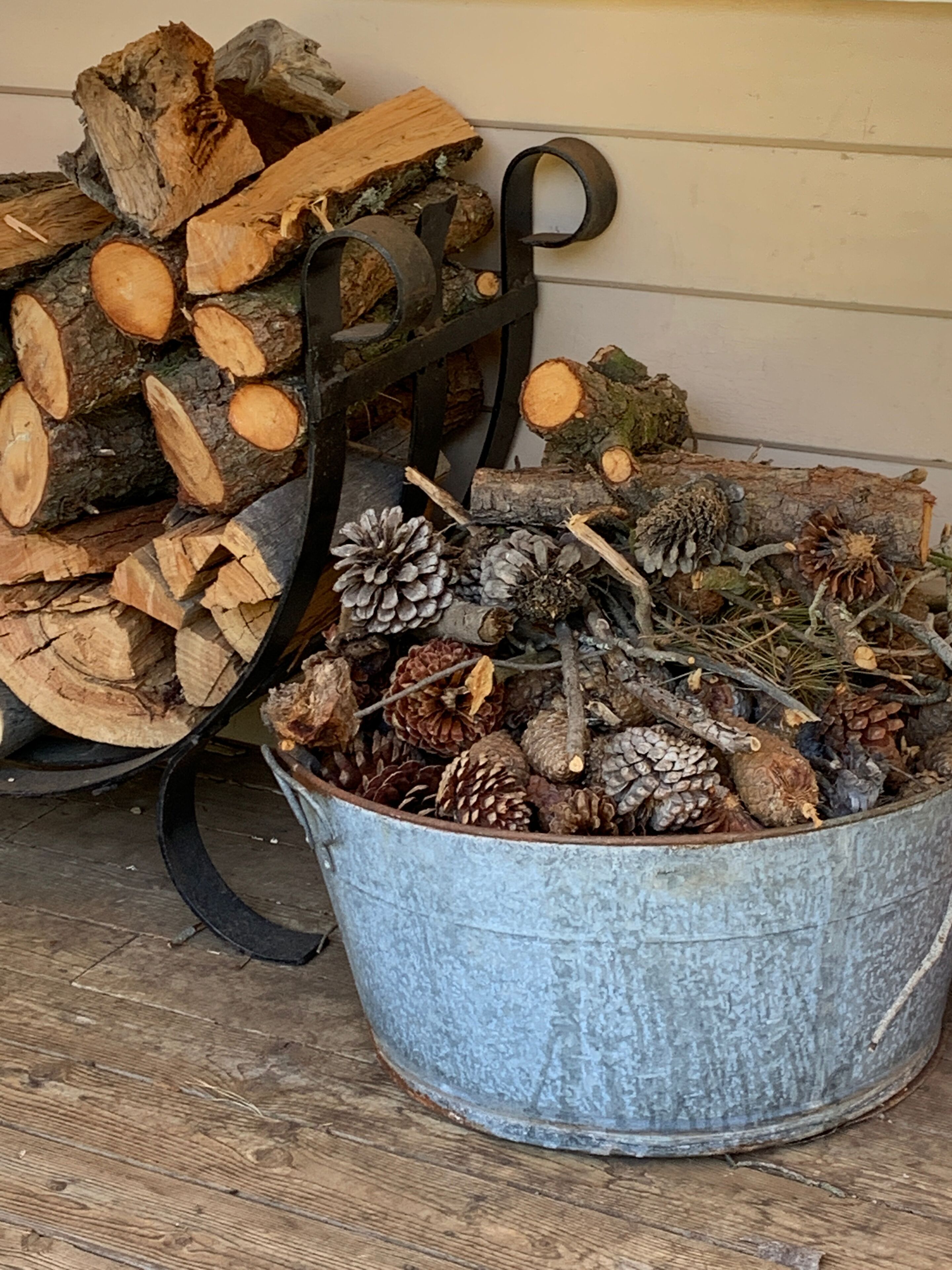 A metal bucket filled with pinecones and branches, placed on a wooden floor next to a stack of firewood.