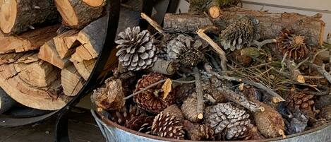 A metal bucket filled with pinecones and branches, placed on a wooden floor next to a stack of firewood.