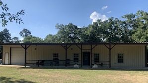 A single-story building with a large garage door, a covered porch with benches, and a grassy lawn. - PERFECTLY CUTE COUNTRY BARDOMINIUM ON 11 ACRES (Bedias)