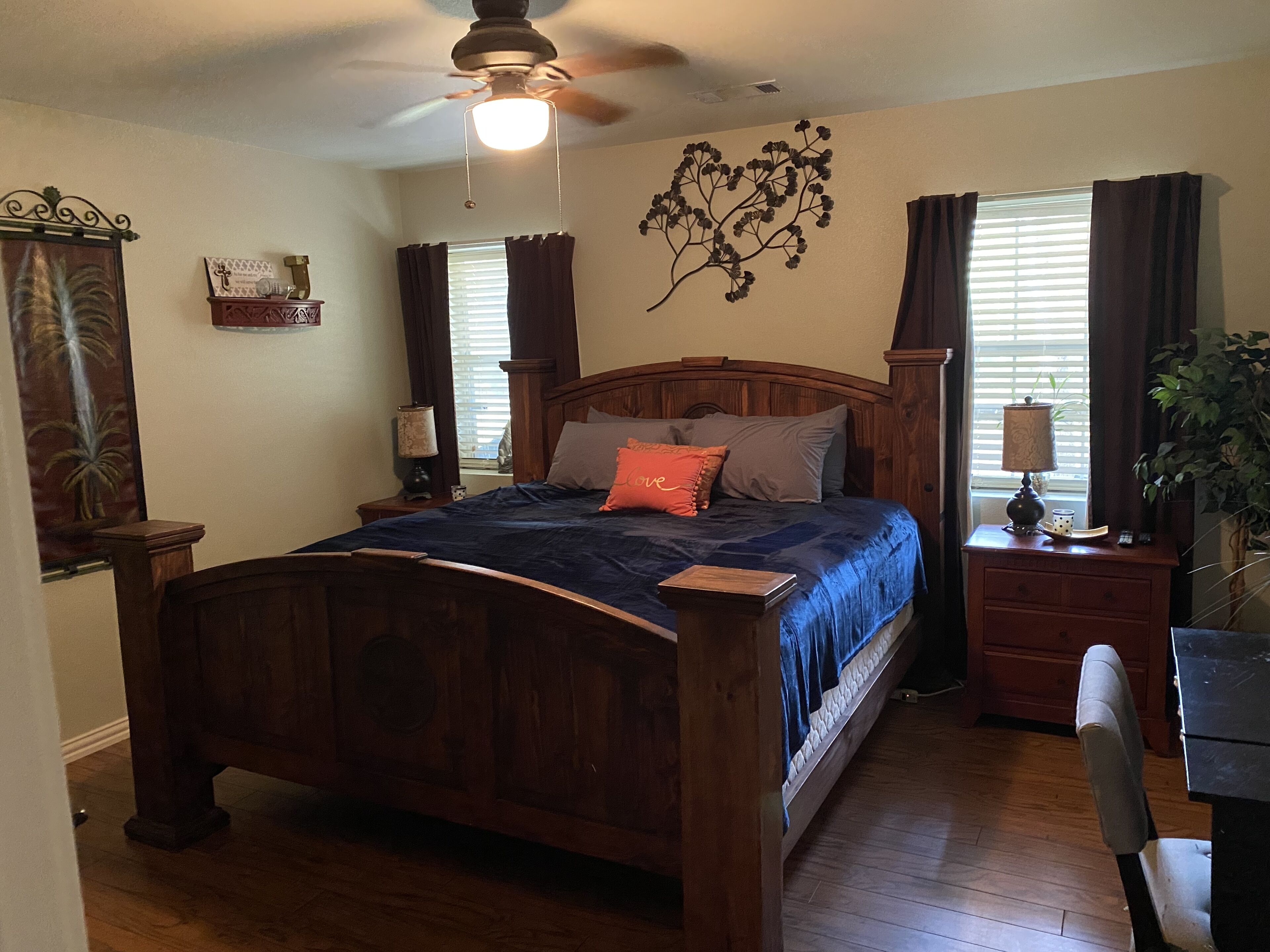 A bedroom with a wooden bed, a nightstand with a lamp, a dresser, a chair, and a ceiling fan.