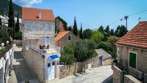 Un bâtiment en pierre avec un toit recouvert de tuiles rouges, un balcon doté de sièges extérieurs et une petite structure adjacente surmontée d’une auvent bleue.