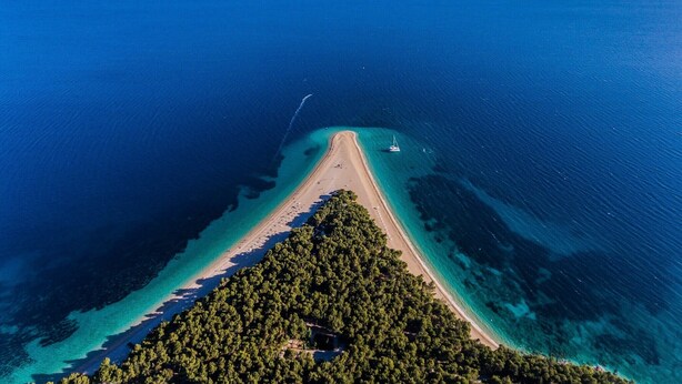 Vue aérienne d’une zone côtière avec une plage étroite, des eaux d’un bleu limpide et une péninsule boisée.