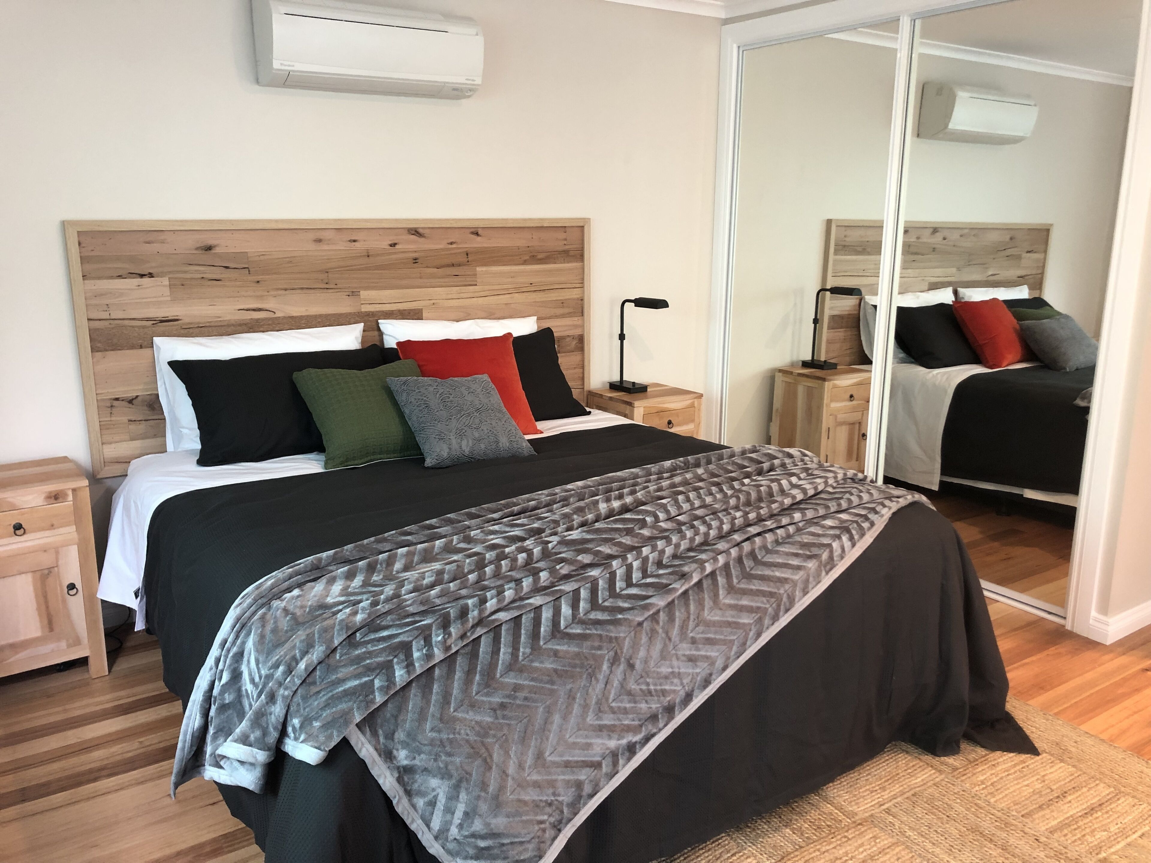 A neatly arranged bedroom with a wooden headboard, a large bed with multiple pillows, a bedside table with a lamp, and a mirror reflecting the room.