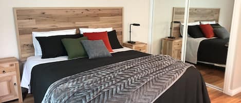 A neatly arranged bedroom with a wooden headboard, a large bed with multiple pillows, a bedside table with a lamp, and a mirror reflecting the room.