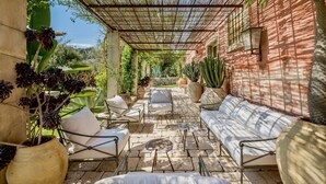 A covered outdoor patio with stone flooring, white cushioned seating, and various potted plants.