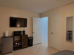Una habitación con televisión de pantalla plana, refrigerador pequeño, microondas, cafetera, basurero y cama.