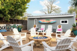 An outdoor seating area with white Adirondack chairs, a fire pit, and a building with a sign reading 'The Island'. - The Bung – A Little Slice of Paradise! 🦩🌺 (Lubbock)