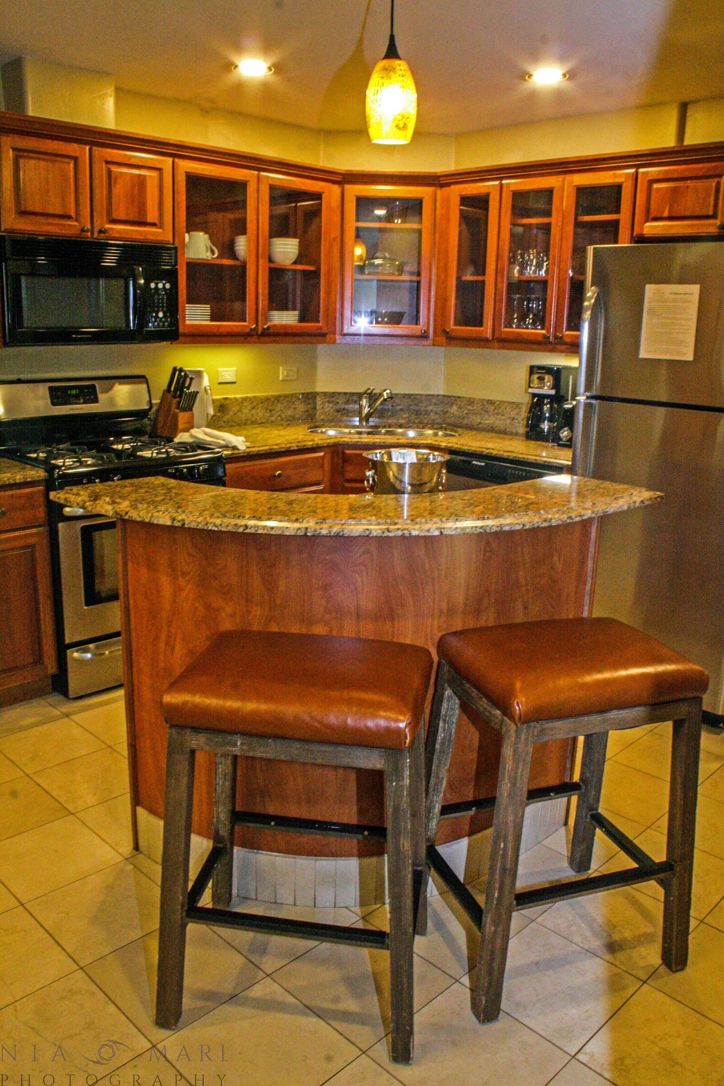 A kitchen with wooden cabinets, a granite countertop, a stainless steel refrigerator, and two leather bar stools.