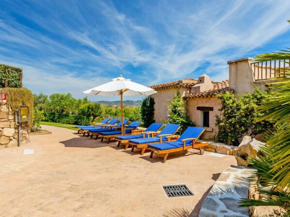 Un patio con sillones de sol y una sombrilla blanca, rodeado de vegetación y un muro de piedra.