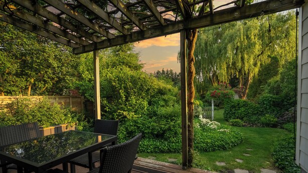 An outdoor patio with a glass table and chairs, surrounded by lush greenery and a wooden pergola. - Secret Garden Cottage in Old Town Niagara-on-the-Lake (Niagara-on-the-Lake)