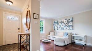 A modern living room with hardwood flooring, a white sofa, a round coffee table, and a large floral painting on the wall. - Secret Garden Cottage in Old Town Niagara-on-the-Lake (Niagara-on-the-Lake)