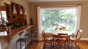 A dining area with a wooden table and chairs, a kitchen counter with stools, and a large window with a view of greenery. - Harmony House - a serene place to relax and renew (Sebastopol)