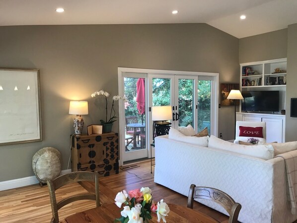 Una sala de estar con un sofá, una mesa de comedor con sillas, un televisor y una estantería. Hay una puerta corrediza de vidrio que da a un área exterior con una mesa y sillas.